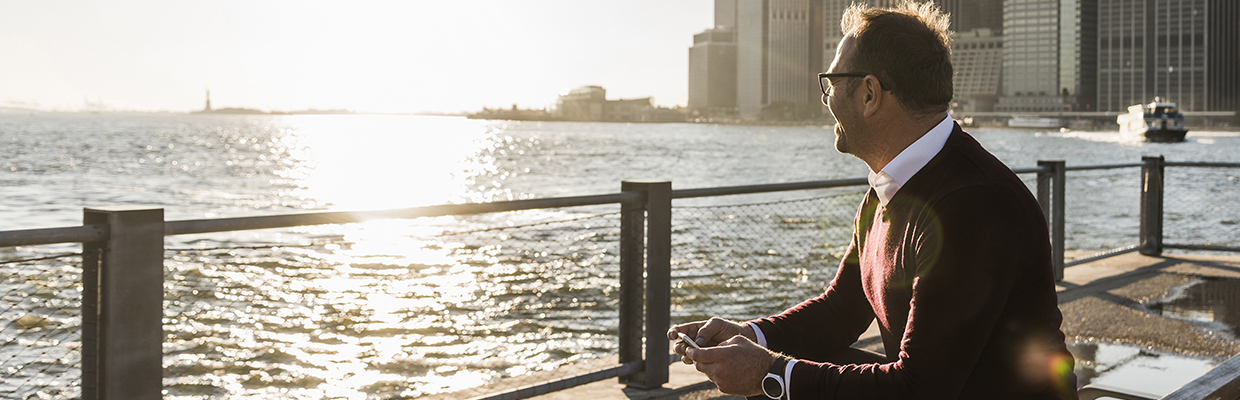 Man in a sweater sitting on a bench with a phone in his hand, looking out over a harbour. Image used for HSBC Malaysia Wealth Solutions page.