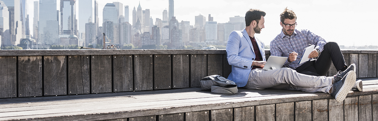 Two guys sitting on a pier having a coffee and comparing notes. One has a laptop, while the other is holding a notebook. Image used for HSBC Malaysia Managing your money page.