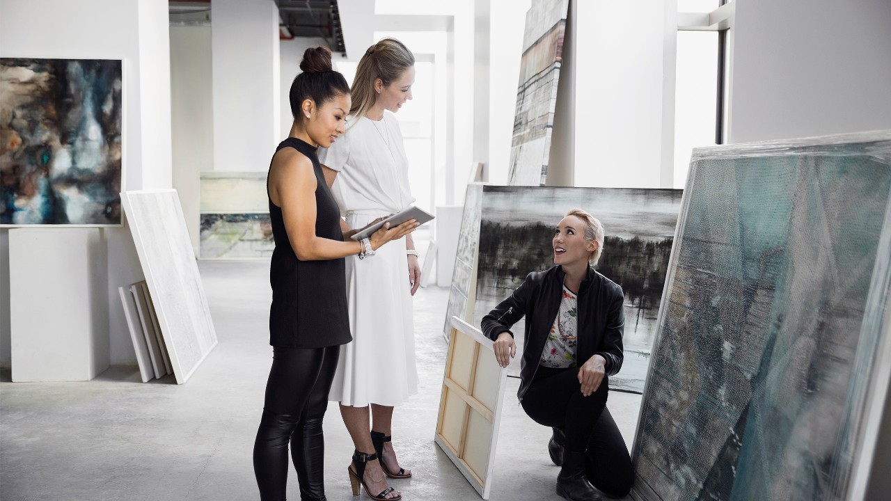 Two women looking at art while the painter explains what they are looking at. Image used for HSBC Malaysia investment page.