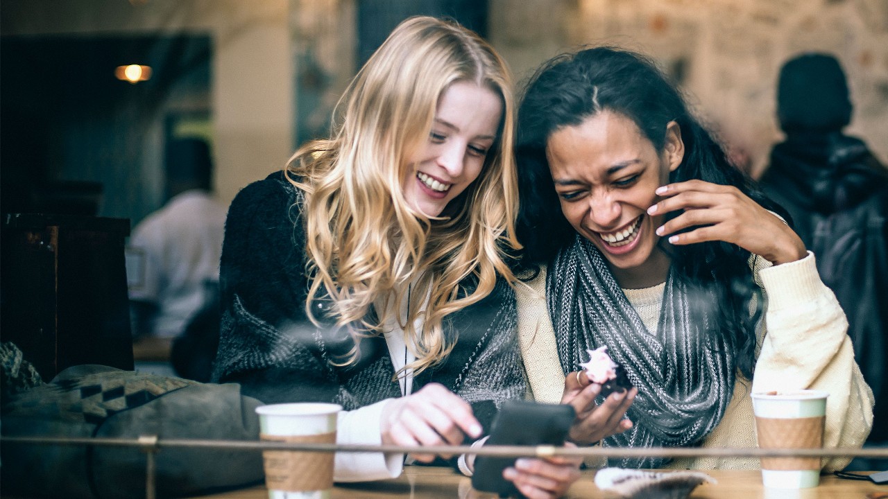 Two friends laughing at something on their phone while having coffee. Image used for HSBC Malaysia Visa debit card page.
