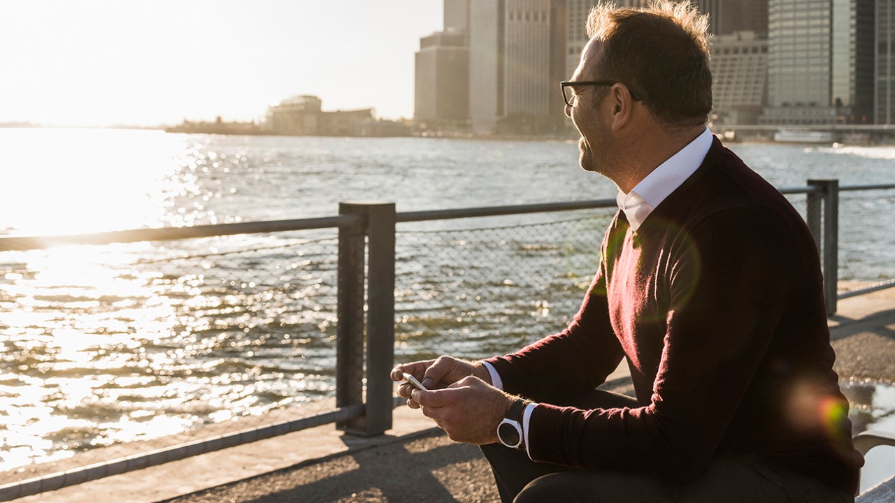 Man in a sweater sitting on a bench with a phone in his hand, looking out over a harbour. Image used for HSBC Malaysia Wealth Solutions page.