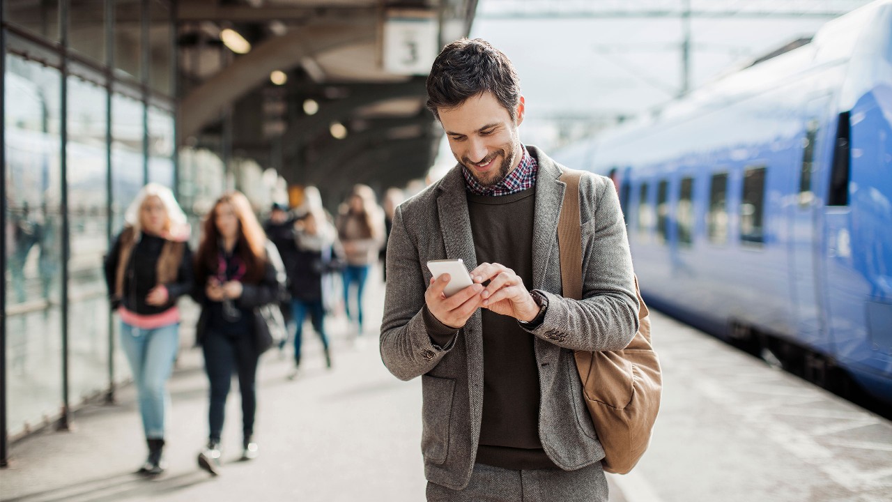 Man in a grey jacket looking at this phone wile standing on a train platform. Image used for HSBC Malaysia bank in your mobile page.