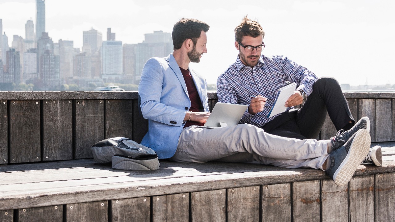 Two guys sitting on a pier having a coffee and comparing notes. One has a laptop, while the other is holding a notebook. Image used for HSBC Malaysia Move money page.